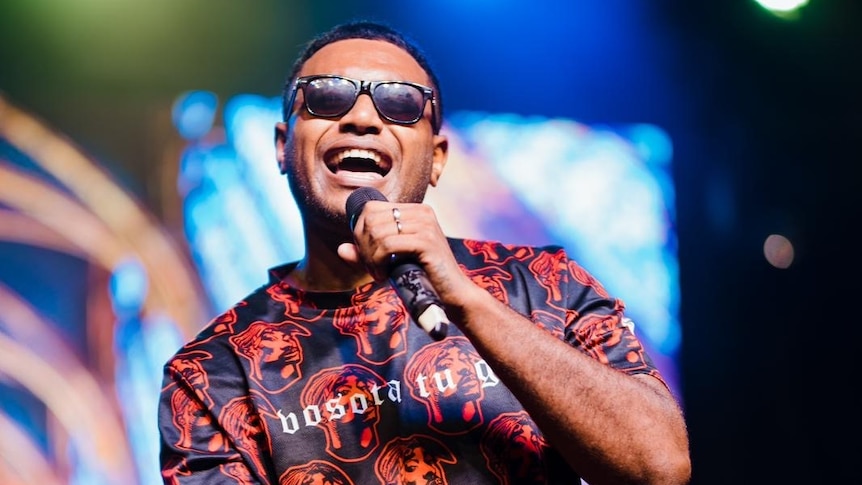 A man wearing sunglasses holds a microphone, performing onstage.