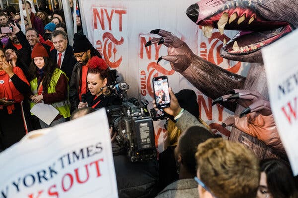 A group of people are tightly gathered in front of a white sign that reads “NYT Guild” and an inflatable rat.