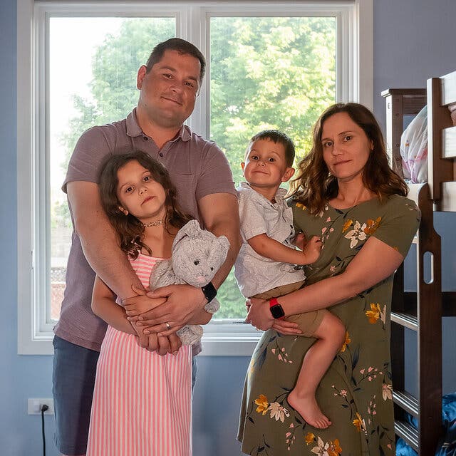 A man and a woman pose with their two children, including a little girl who is wearing a pink dress and holding a stuffed animal and a little boy who is grinning at the camera. In the background is a child’s bunk bed. 
