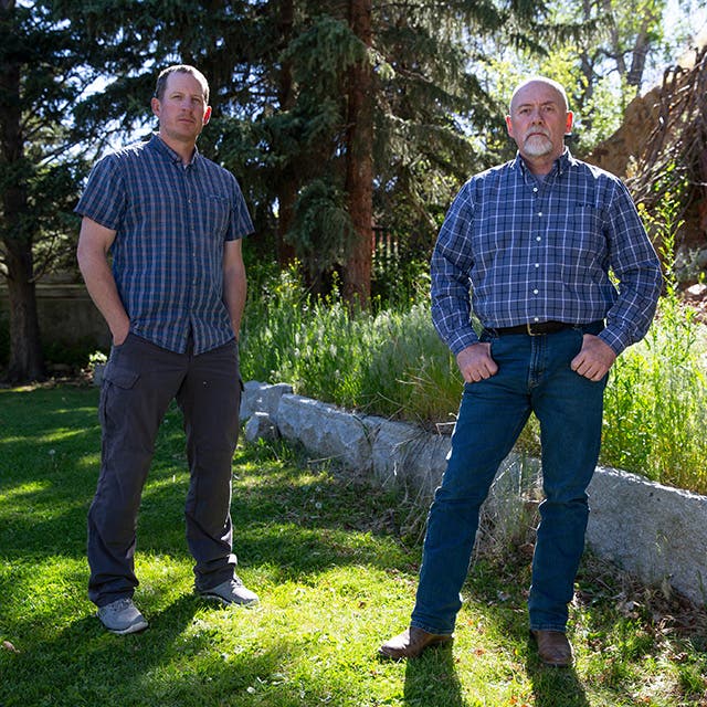 Two men outside in a wooded area. Both are wearing dark pants and a blue plaid shirt.