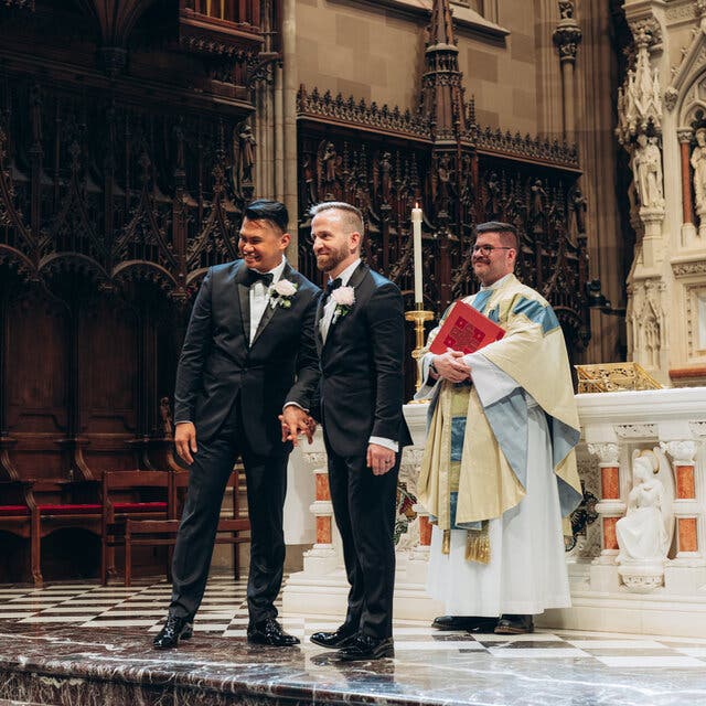 Two smiling grooms in tuxedos, with a pale pink rose on their lapels, stand at the altar holding hands and facing their guests at Trinity Church Wall Street in New York. Standing behind them is a priest, in an off-white and aqua robe, holding a large red book in both arms across his chest. 
